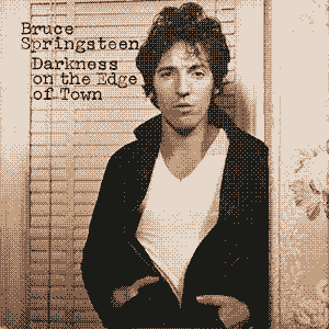 The album cover for Bruce Springsteen's Darnkess on the Edge of Town. The cover is a photograph of Springsteen, who is centered in the frame, wearing a white tee shirt and a black leather jacket. His hands are in the jacket pockets and he is leaning against a wall in a home, looking at the camera with a lazy sort of focus.